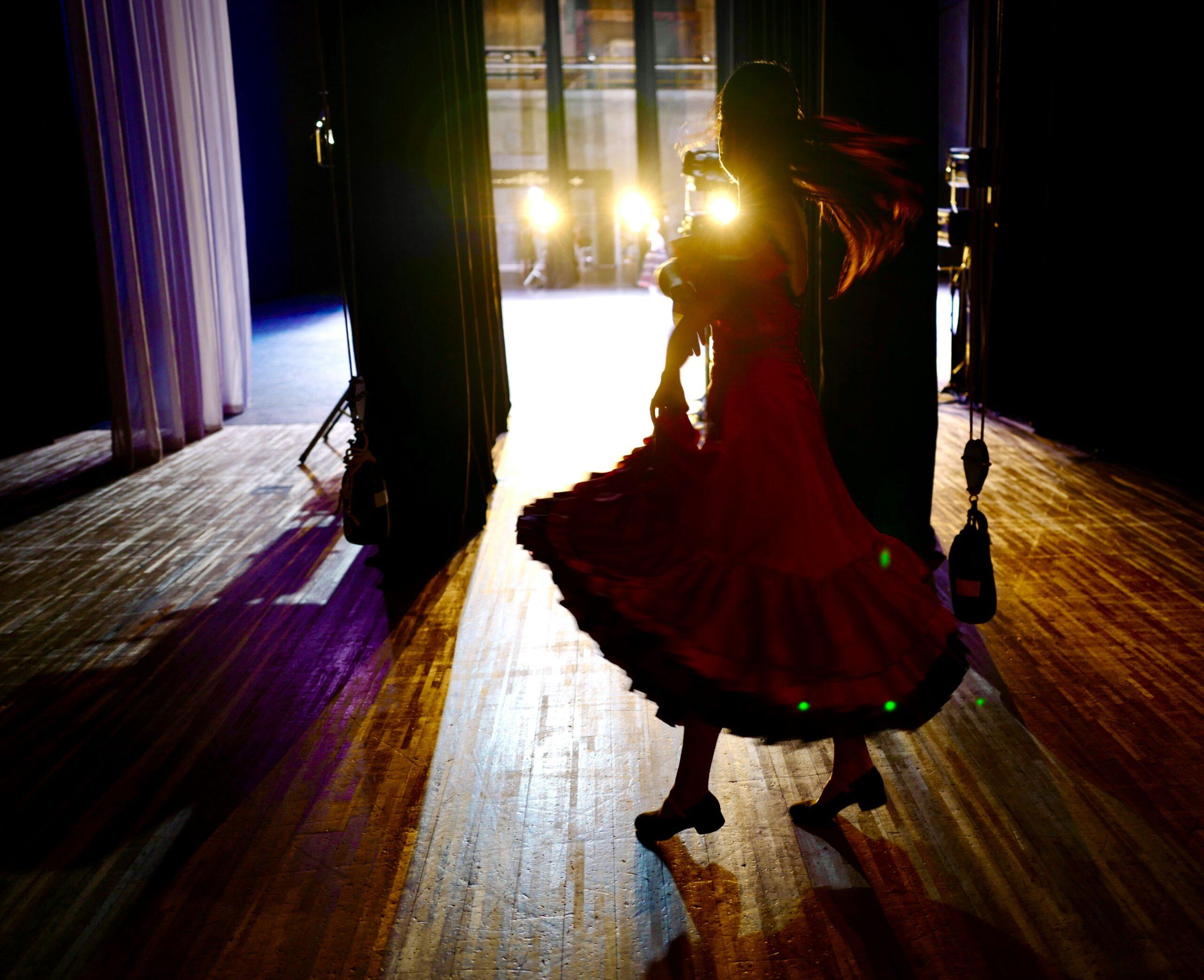 Une femme en robe rouge danse sur scène, illuminée par des projecteurs dans une salle de spectacle
