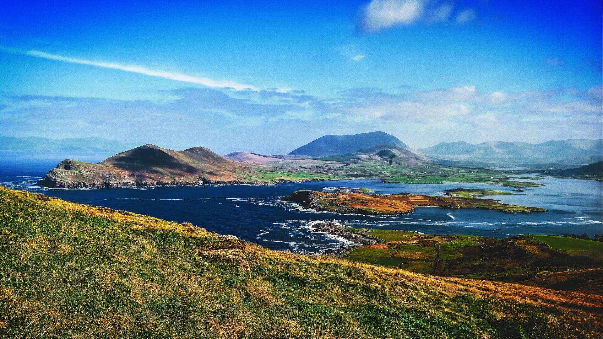 Paysage côtier irlandais avec îles volcaniques, montagnes verdoyantes et baie bleue sous ciel dégagé