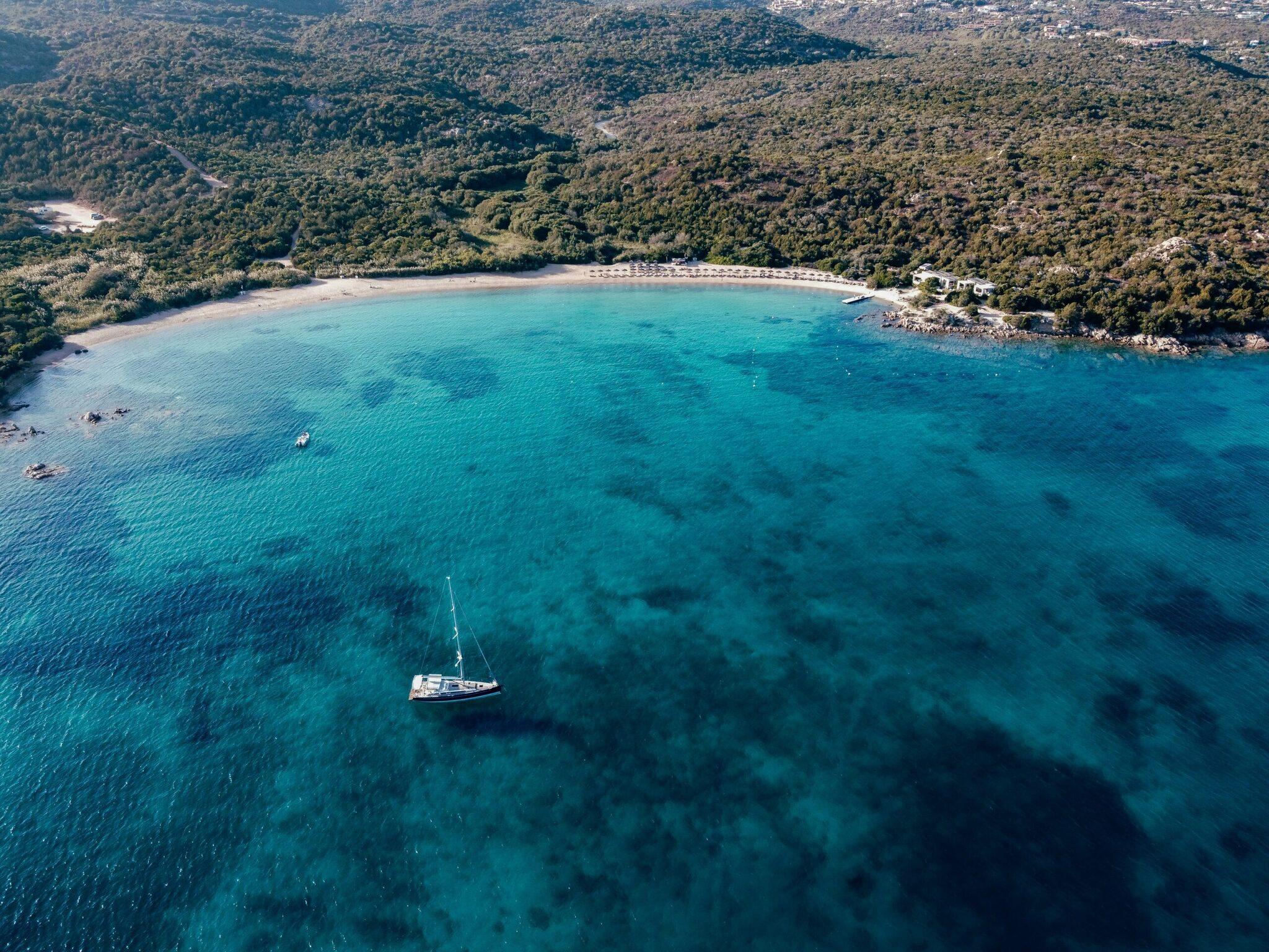 Aerial view of a Mediterranean bay with turquoise waters, a sailing boat, sandy beach and coastal forest