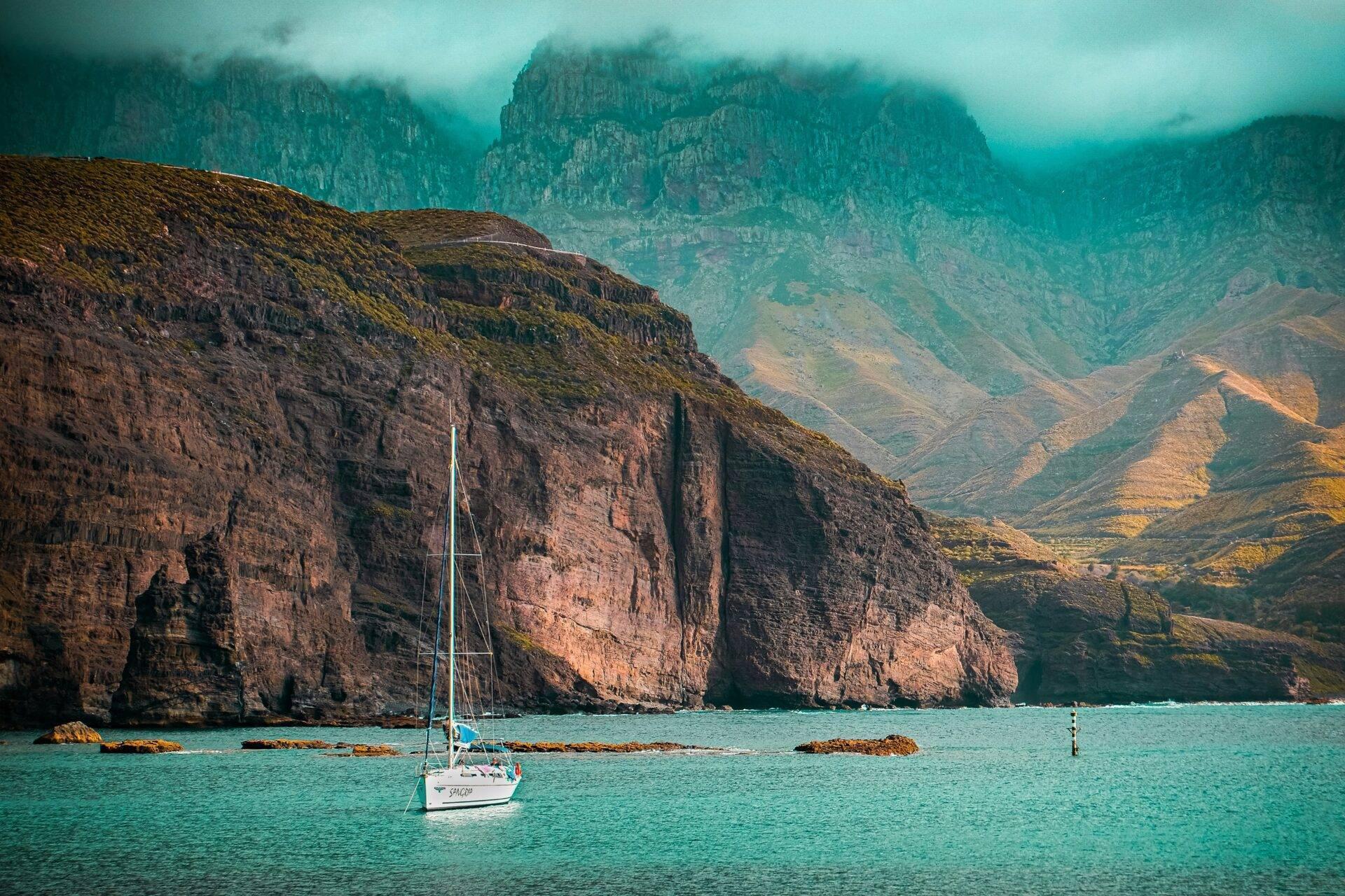 Un voilier blanc ancré dans une baie turquoise encadrée de falaises rocheuses imposantes et de montagnes verdoyantes