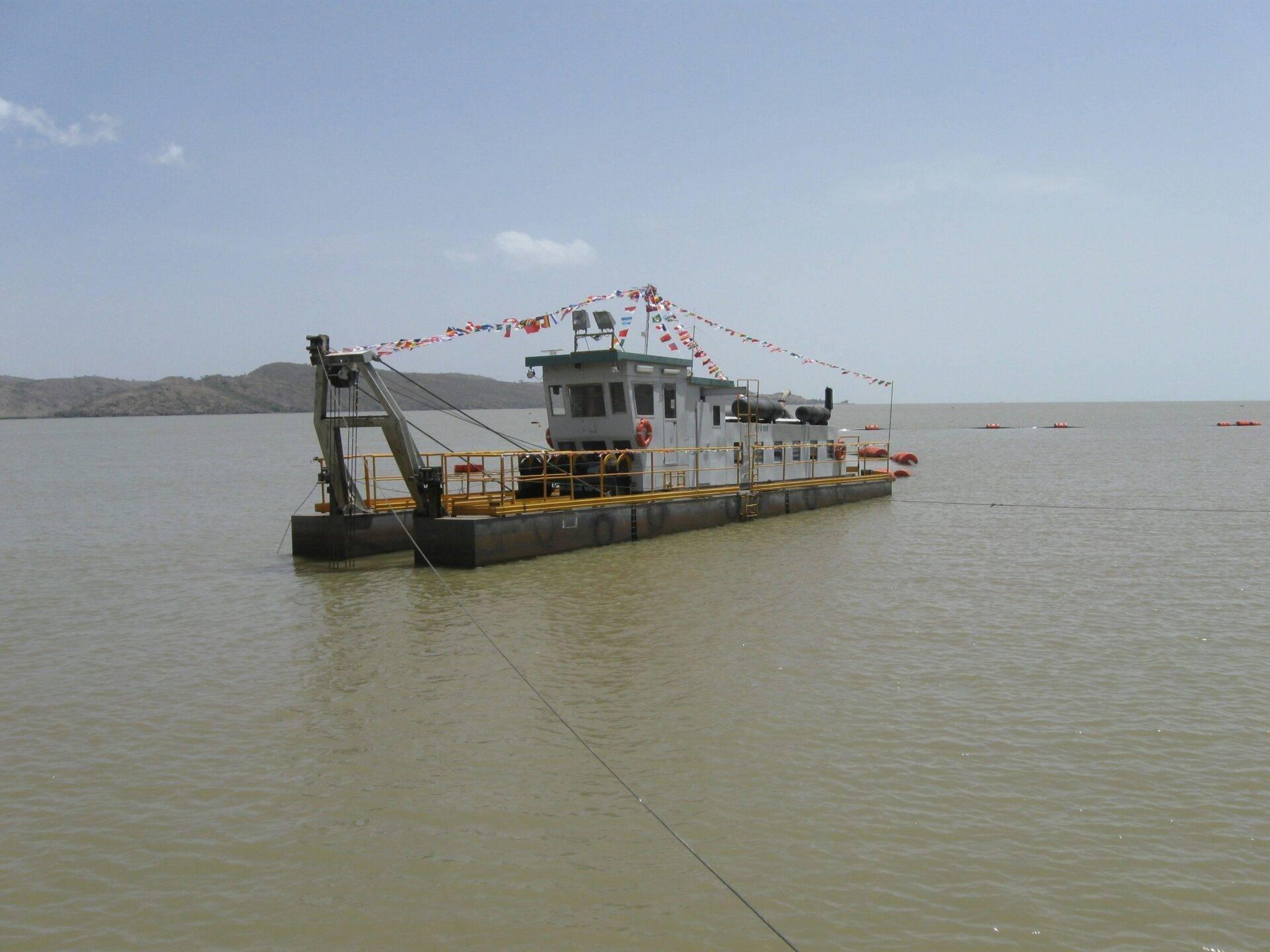 Un bateau de transport fluvial blanc et jaune ancré dans une baie calme, décorée de drapeaux colorés, avec des montagnes en arrière-plan