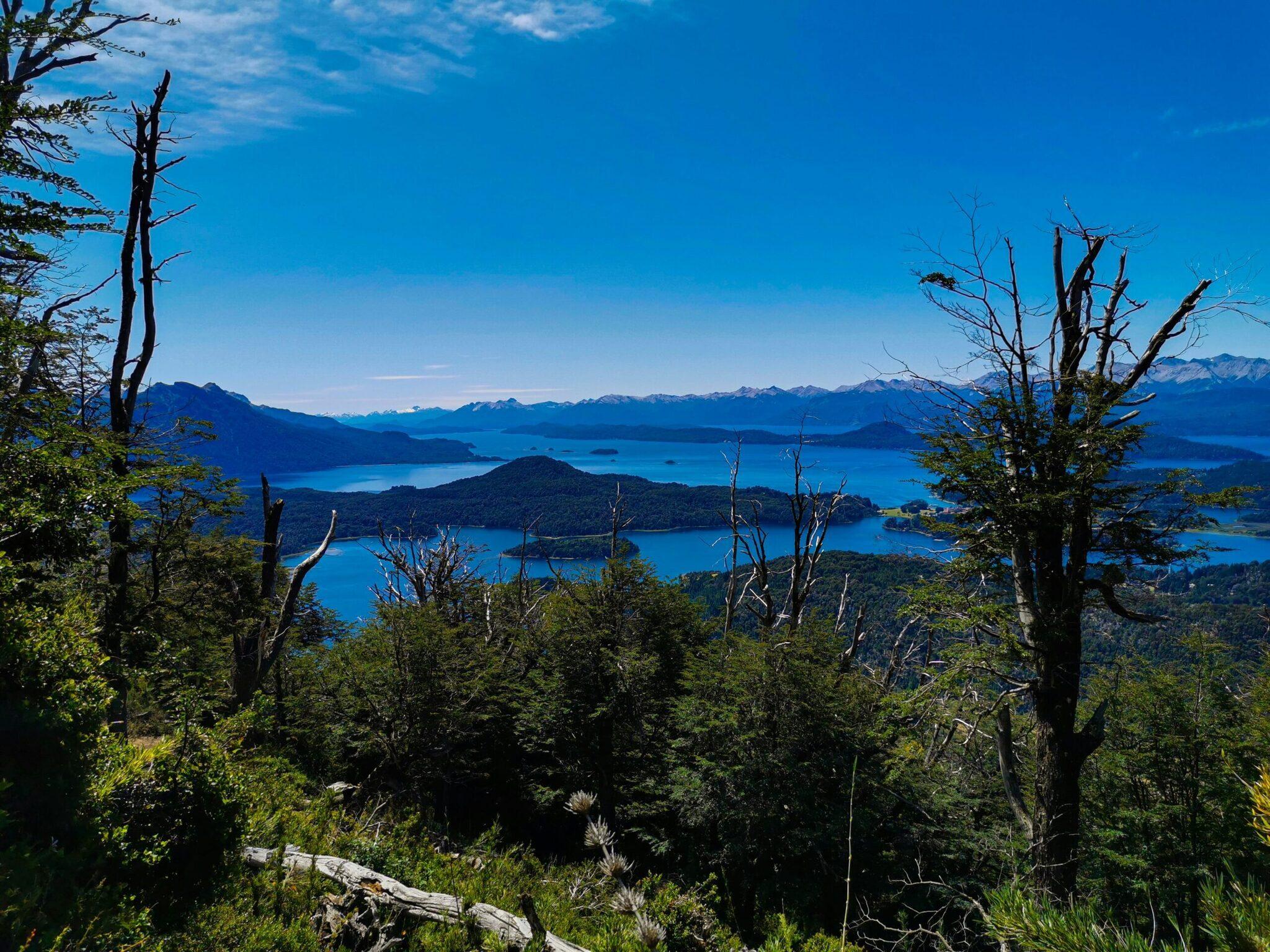 Vue panoramique d’un lac turquoise entouré de forêts denses et de montagnes enneigées sous un ciel dégagé