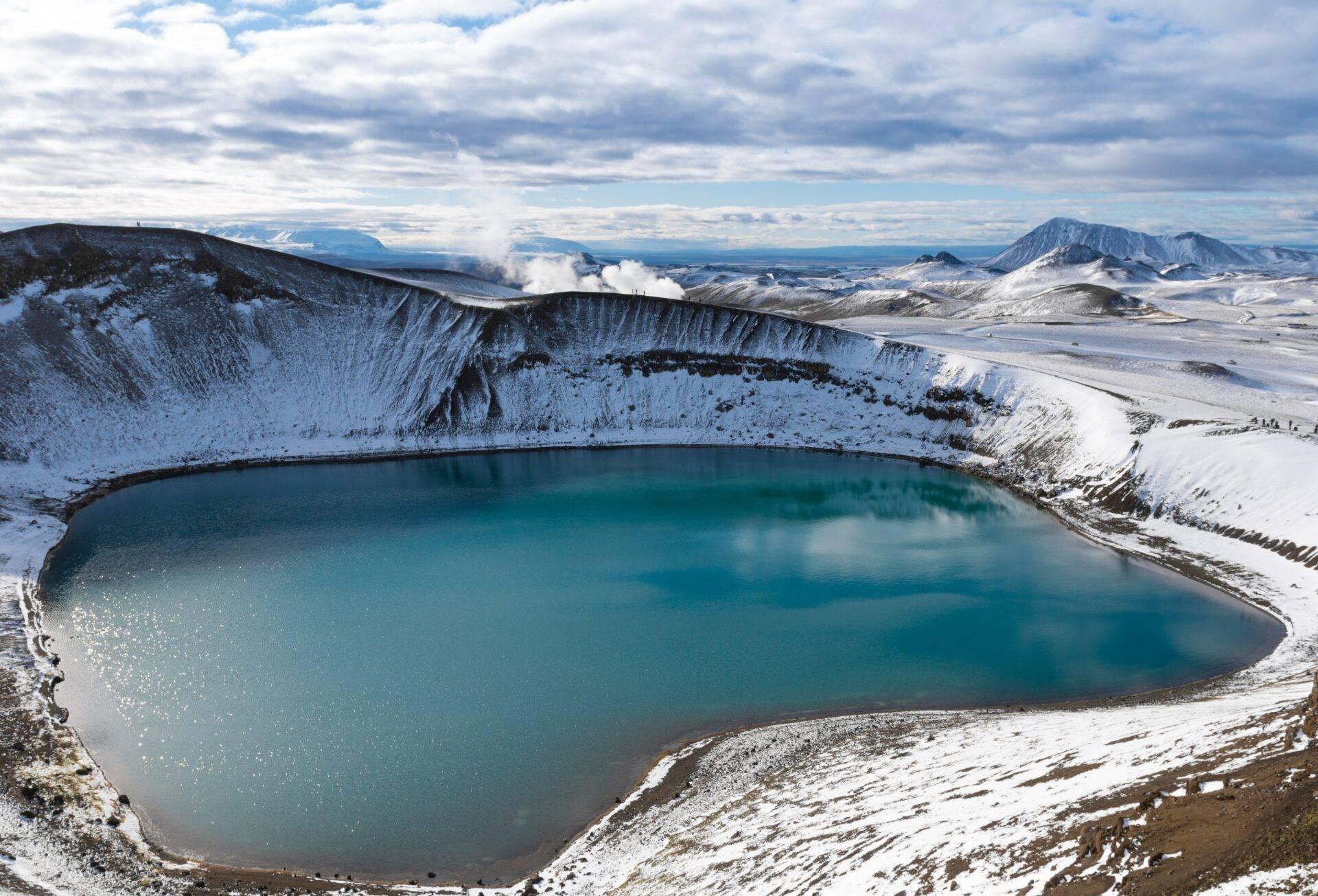 Un cratère volcanique enneigé contenant un lac turquoise bordé de montagnes enneigées sous un ciel nuageux