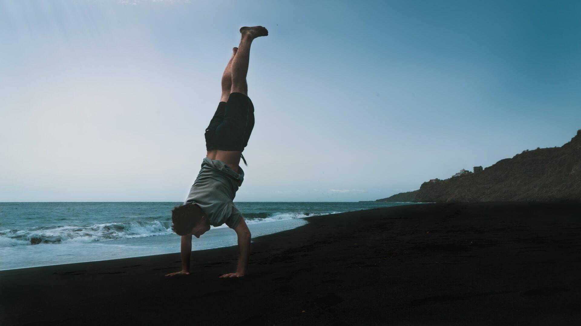 Un homme en équilibre sur les mains sur une plage de sable noir face à l’océan turquoise et des falaises