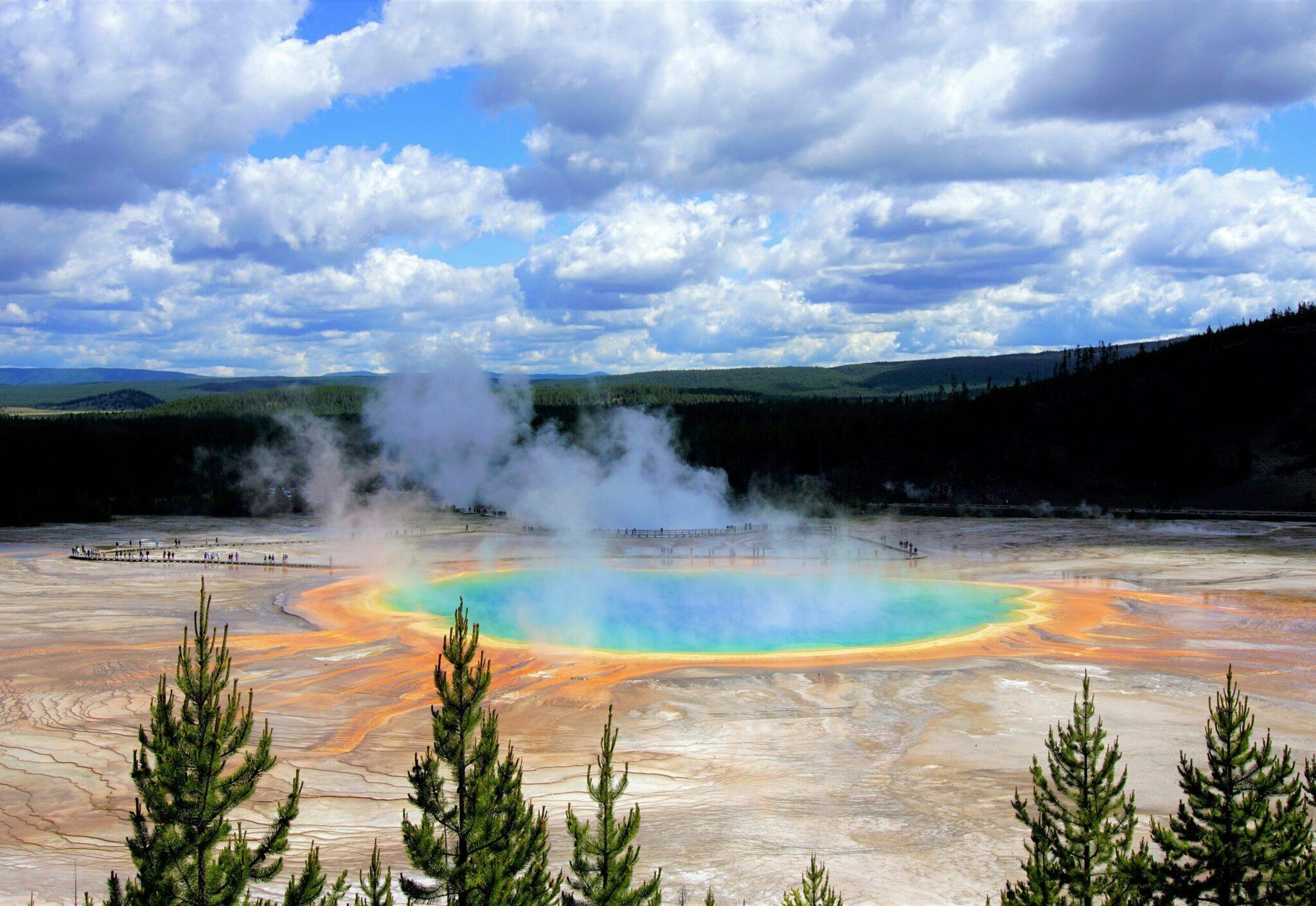 Un geyser coloré aux teintes bleu turquoise et orange émet de la vapeur dans un paysage géothermique forestier sous un ciel nuageux