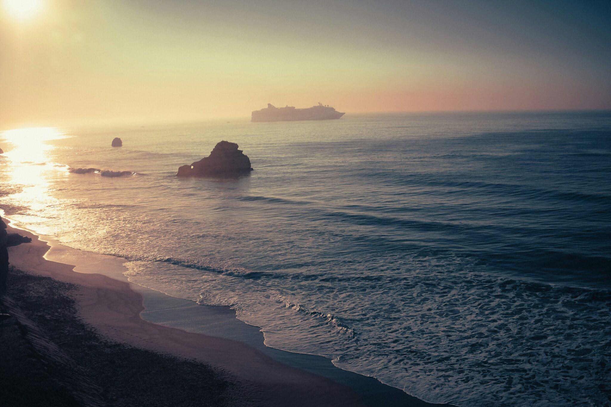 Coucher de soleil doré sur une plage rocheuse avec un navire de croisière à l’horizon
