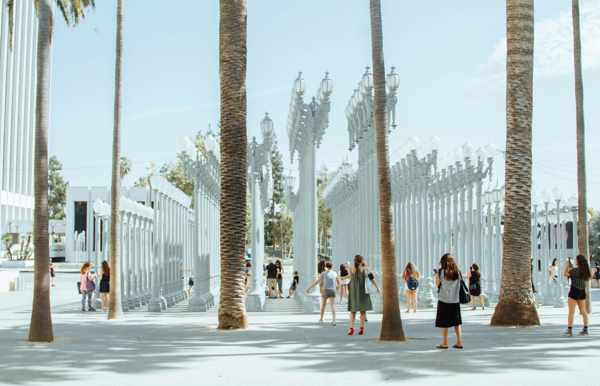Une plaza publique moderne avec colonnes blanches, palmiers et lampadaires, animée par des visiteurs sous un ciel dégagé