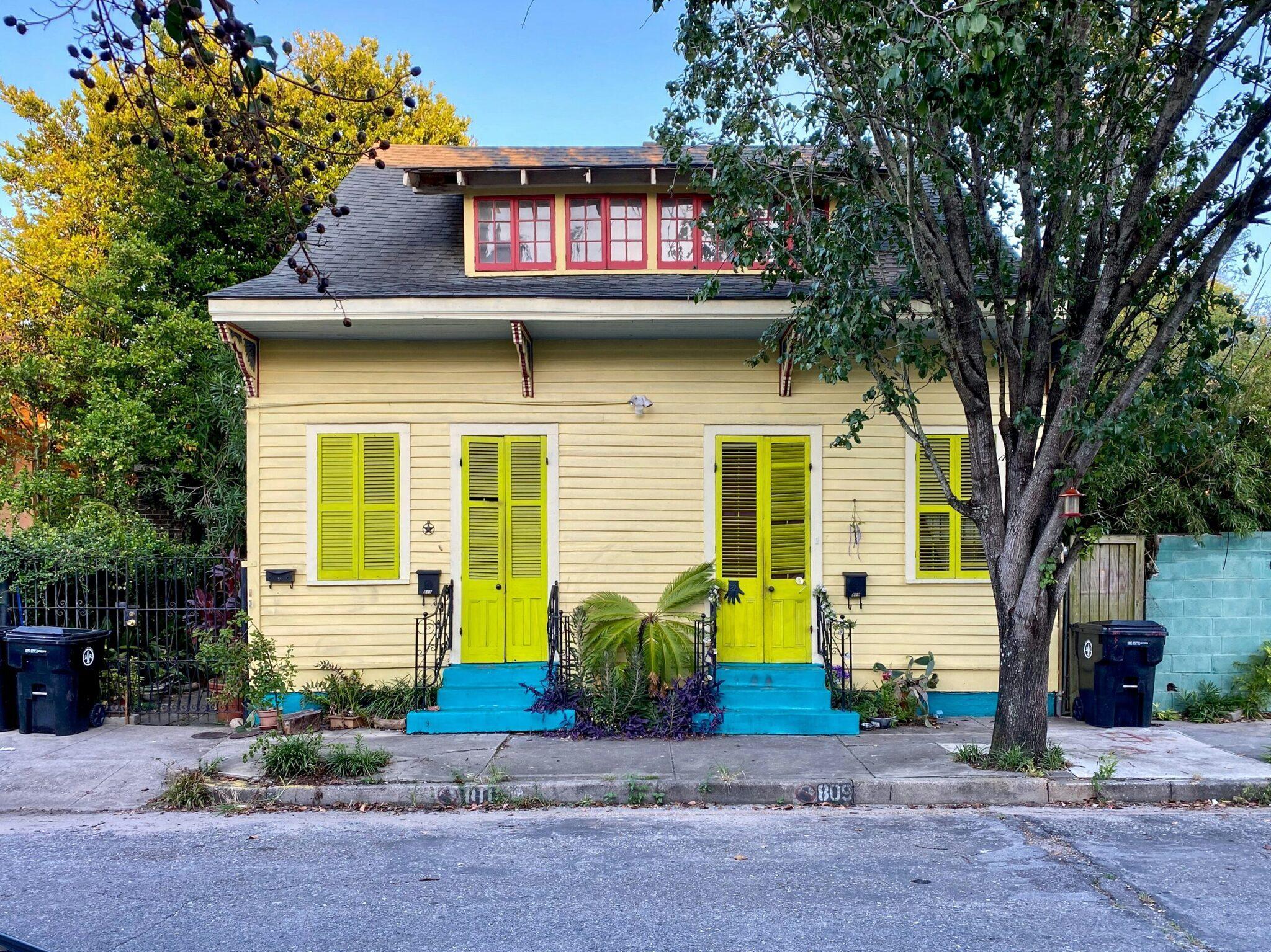 Maison colorée avec portes et volets jaune vif, base bleue et fenêtres rouges à l’étage, entourée de végétation