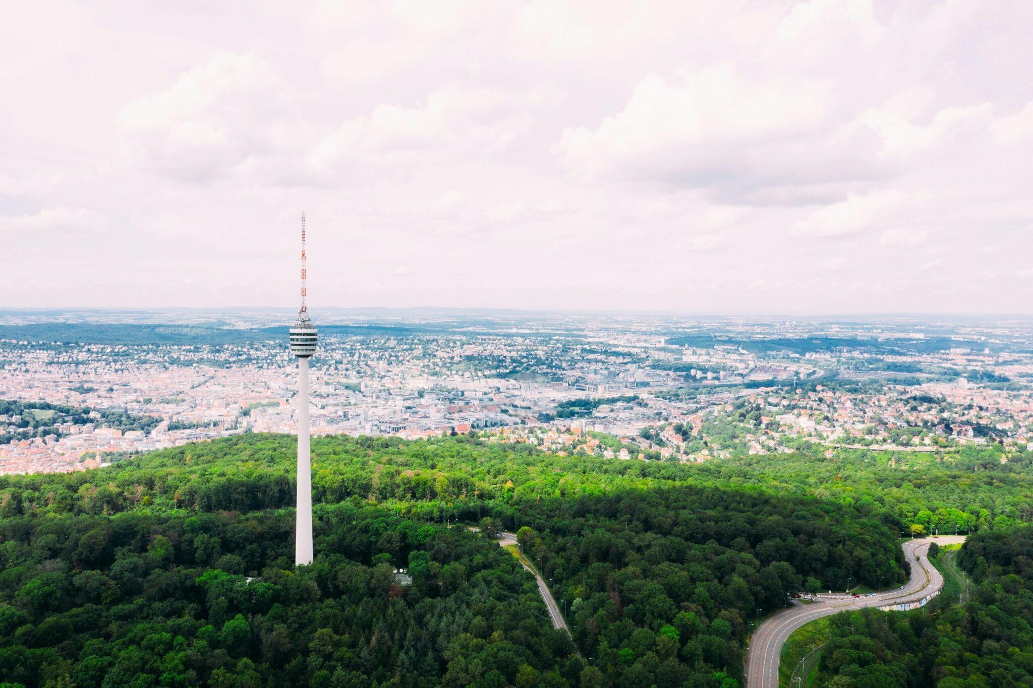 Vue aérienne d’une tour de télévision blanche dominant une forêt dense avec une ville étendue en arrière-plan