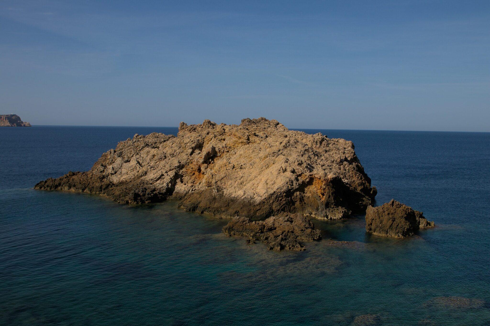 Un îlot rocheux aux formations géologiques spectaculaires émerge de la mer turquoise méditerranéenne sous un ciel bleu dégagé