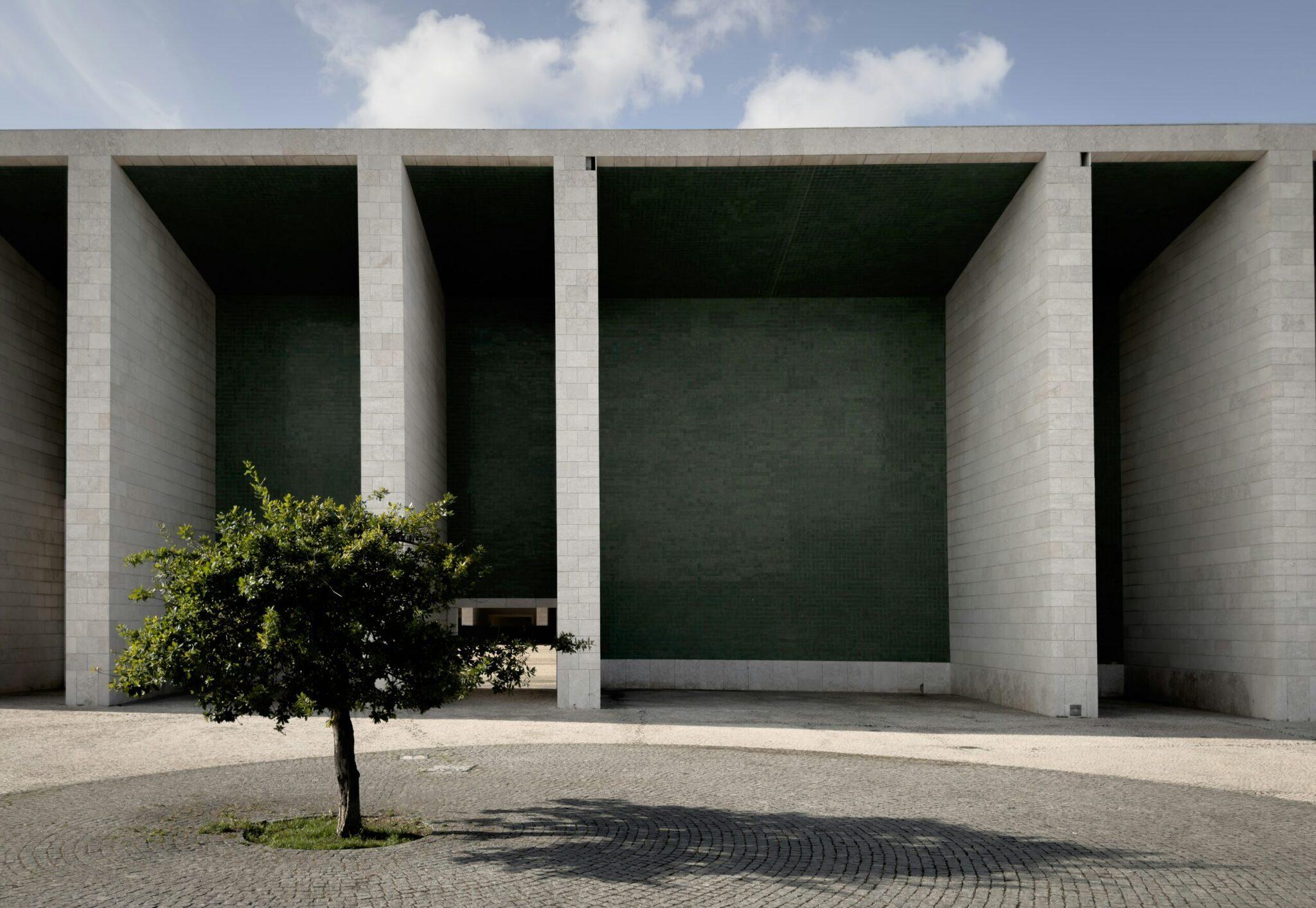 Architecture moderne en béton avec colonnes massives, cour minimaliste et petit arbre, sous ciel dégagé