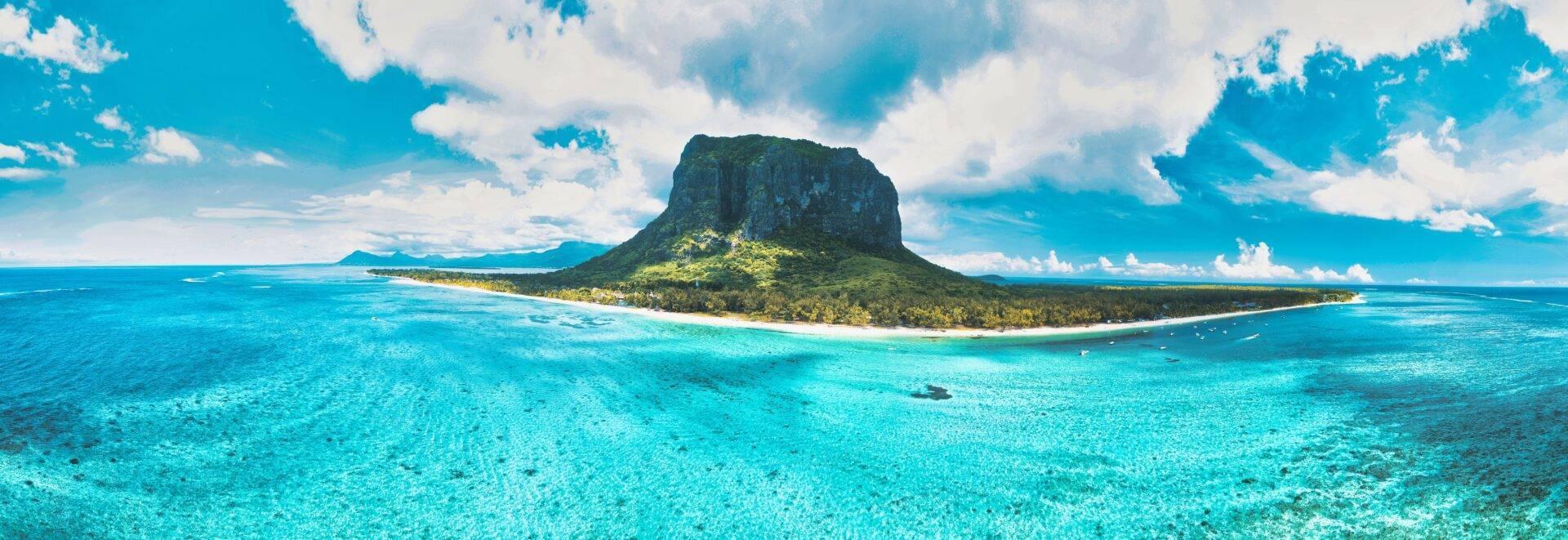 Une île tropicale aux falaises verdoyantes entourée d’eau turquoise cristalline sous un ciel bleu nuageux