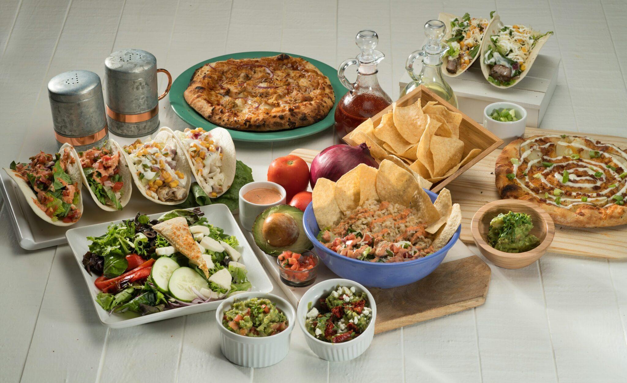 Une table de cuisine blanche présentant un assortiment de cuisine mexicaine avec tacos, nachos, ceviche et salades fraîches