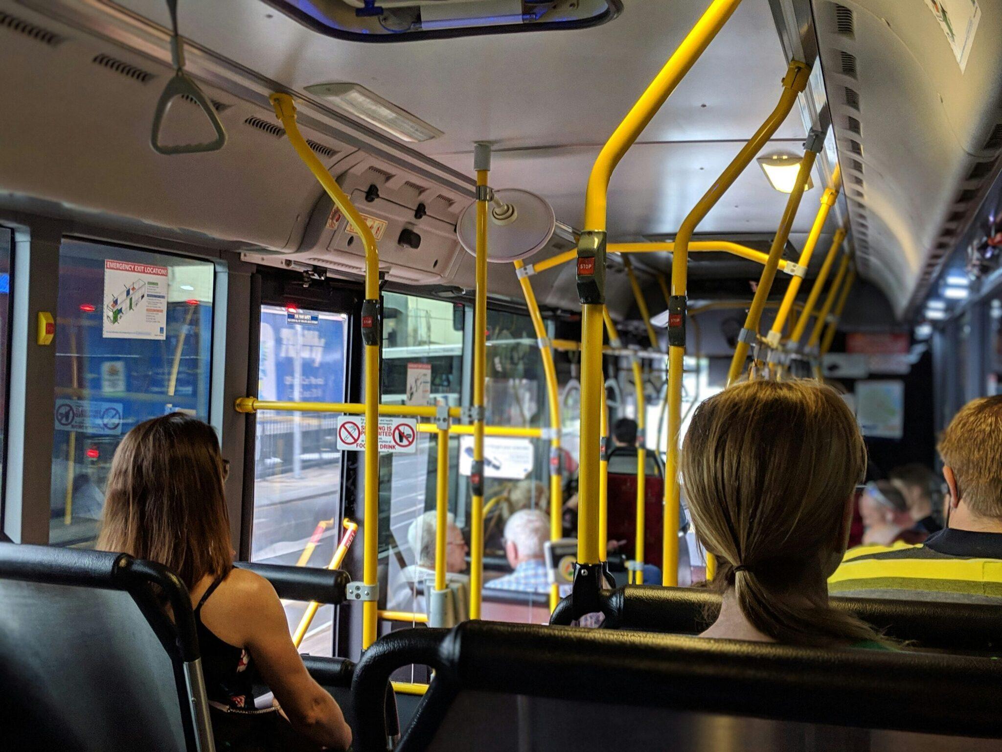 Bus interior with seated passengers, yellow handrails and tinted windows looking out onto the street