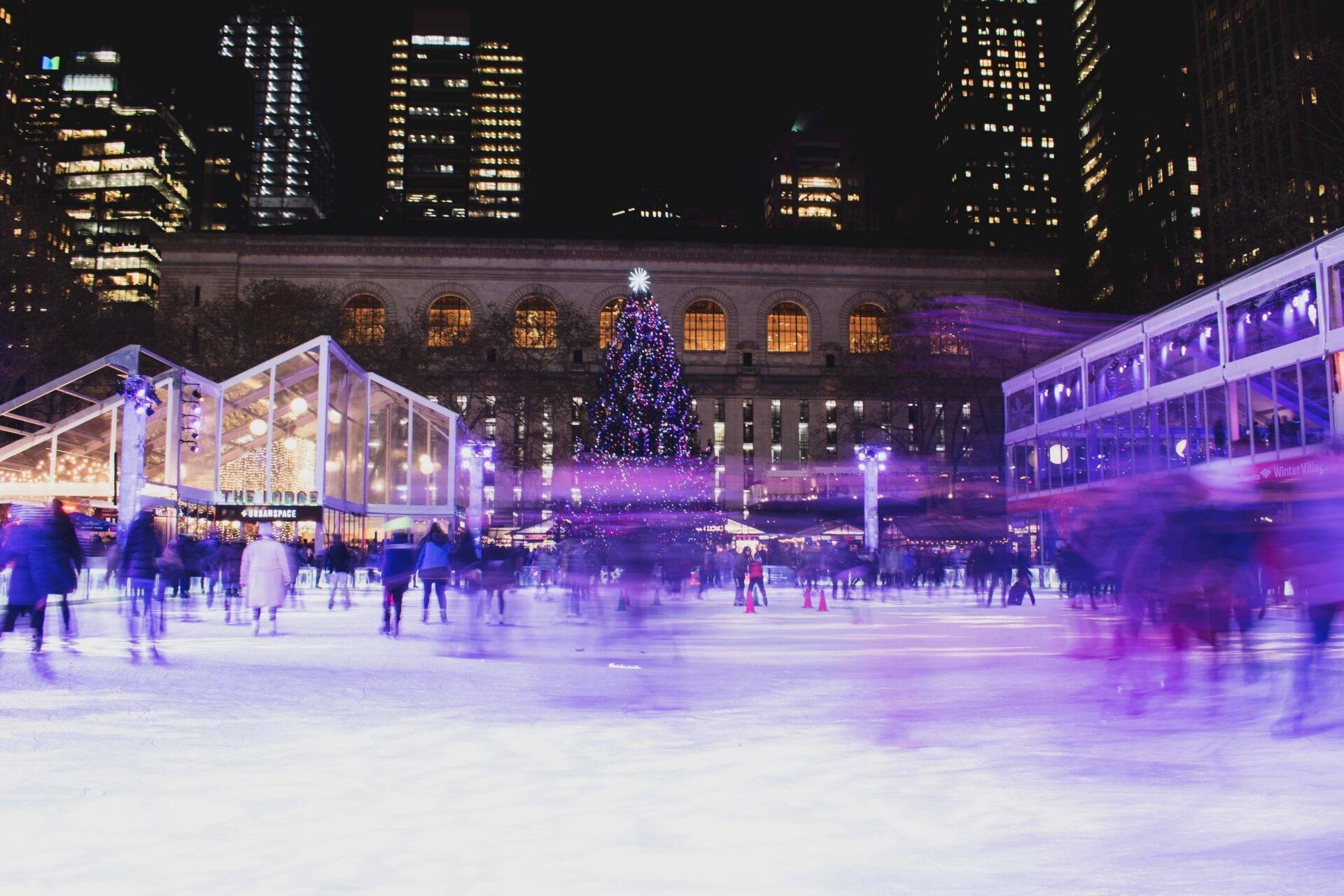Patinoire de Noël illuminée de violet avec sapin décoré et gratte-ciel en arrière-plan, patineurs en mouvement