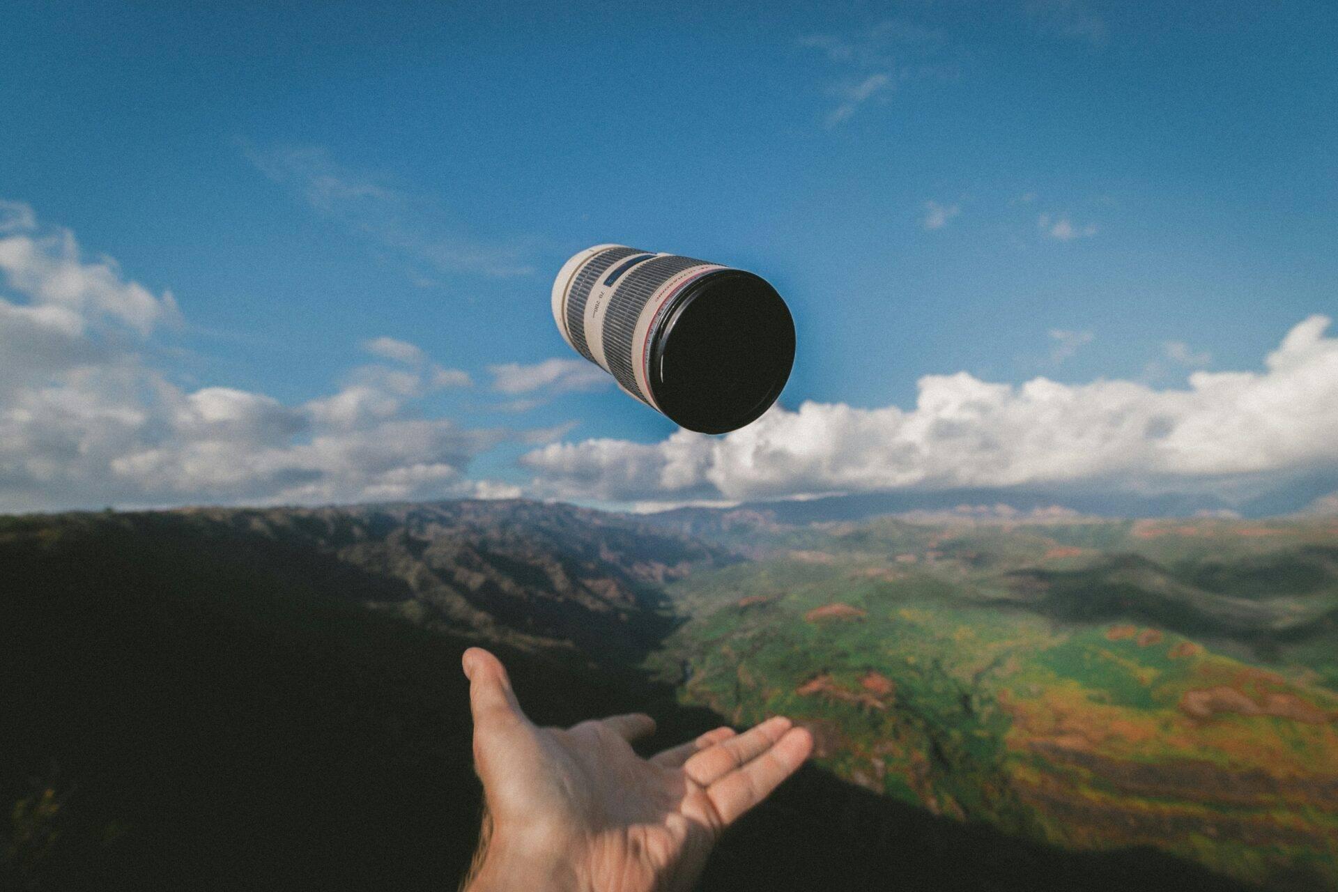 Une main tient un objectif photographique suspendu au-dessus d’un paysage montagneux verdoyant sous un ciel bleu dégagé