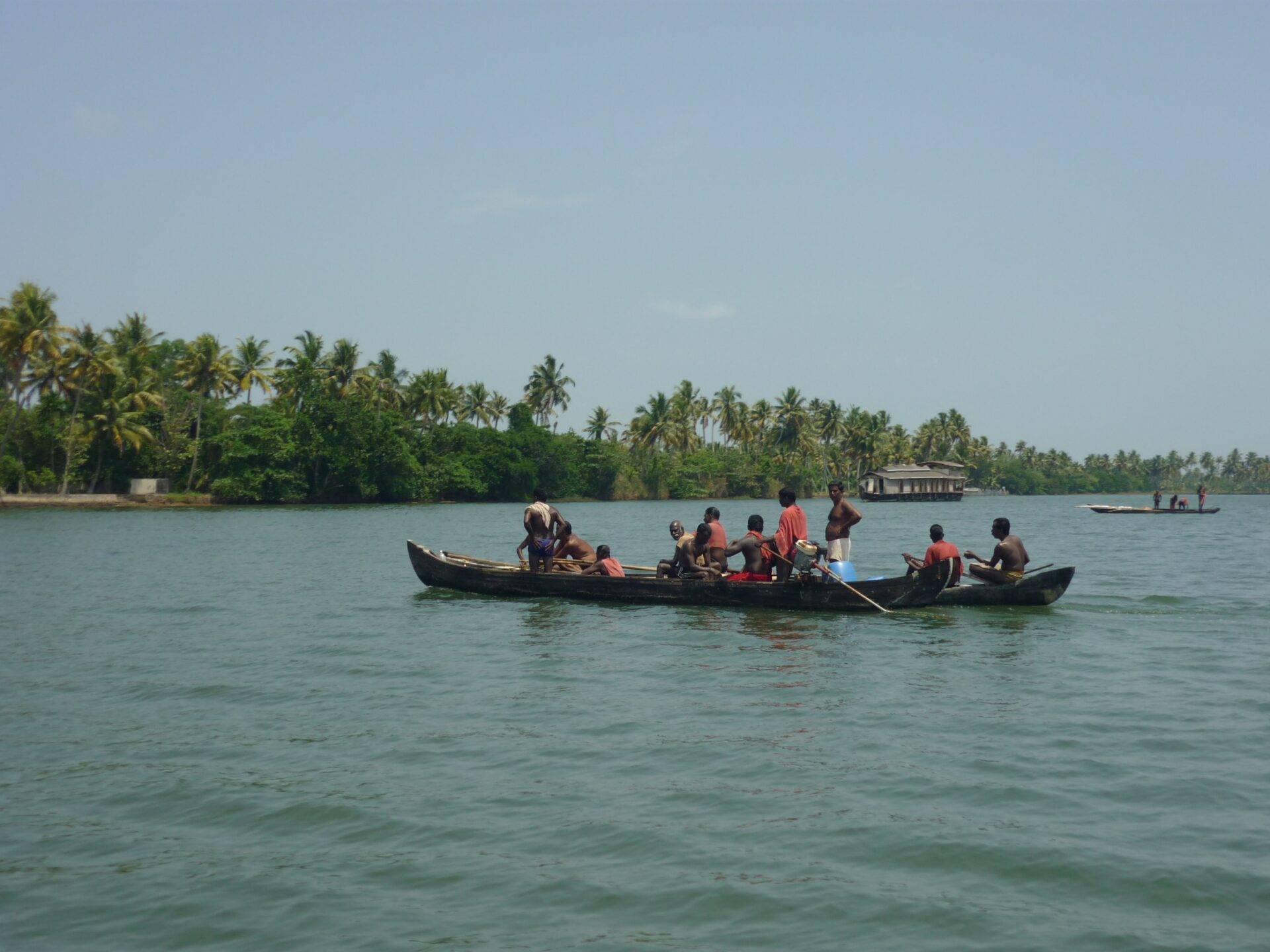 Un bateau traditionnel noir navigue sur des eaux turquoise avec des palmiers et des habitations flottantes en arrière-plan