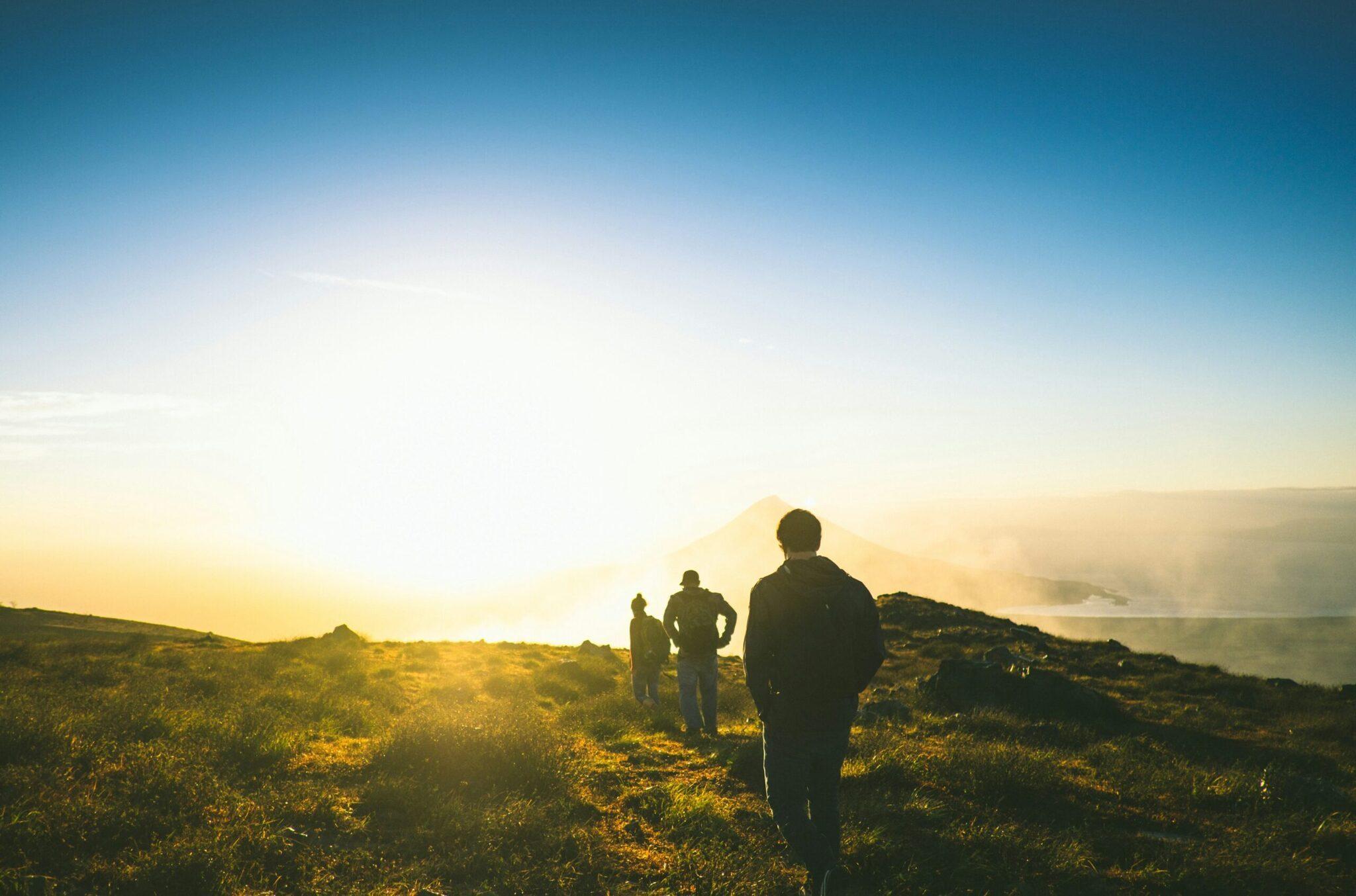Three hikers look out over a snow-capped peak at sunrise from high in a mountain landscape