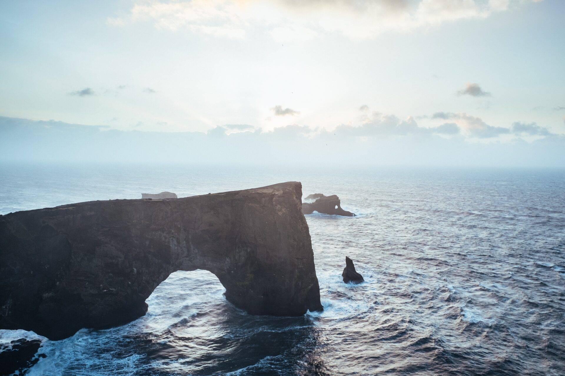 Falaise rocheuse noire avec arche naturelle surplombant l’océan agité sous un ciel nuageux
