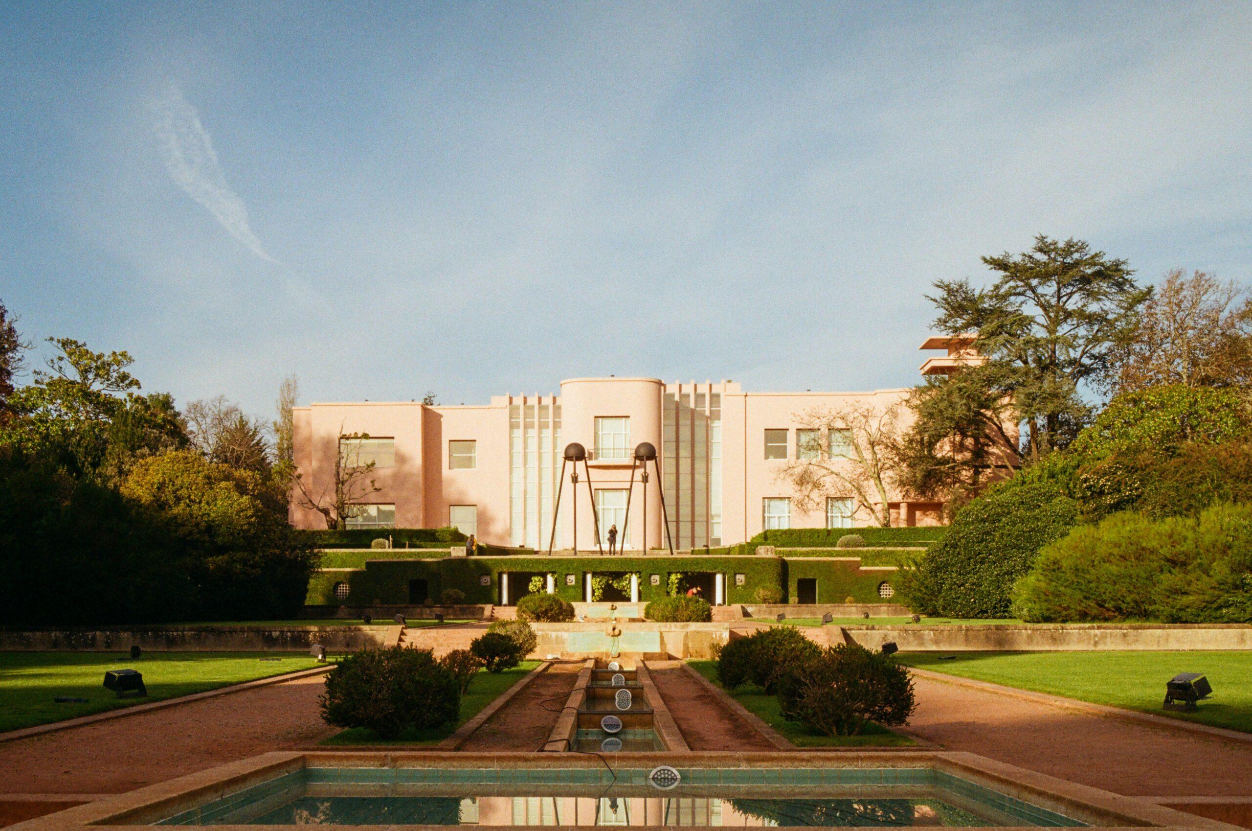 Un bâtiment Art déco rose pâle avec fontaines et bassin de réflexion, entouré de jardins verdoyants et d’arbres
