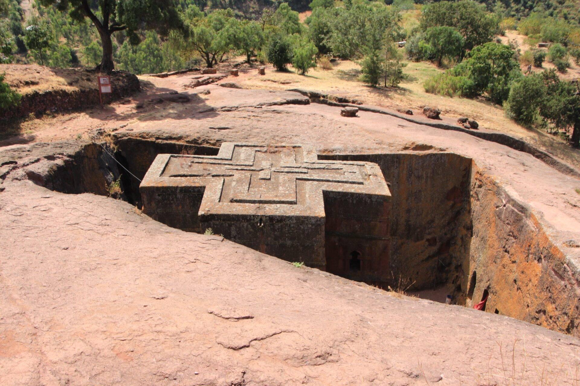 Église rupestre creusée dans la roche avec croix gravée sur le toit, entourée d’arbres et de terrain aride