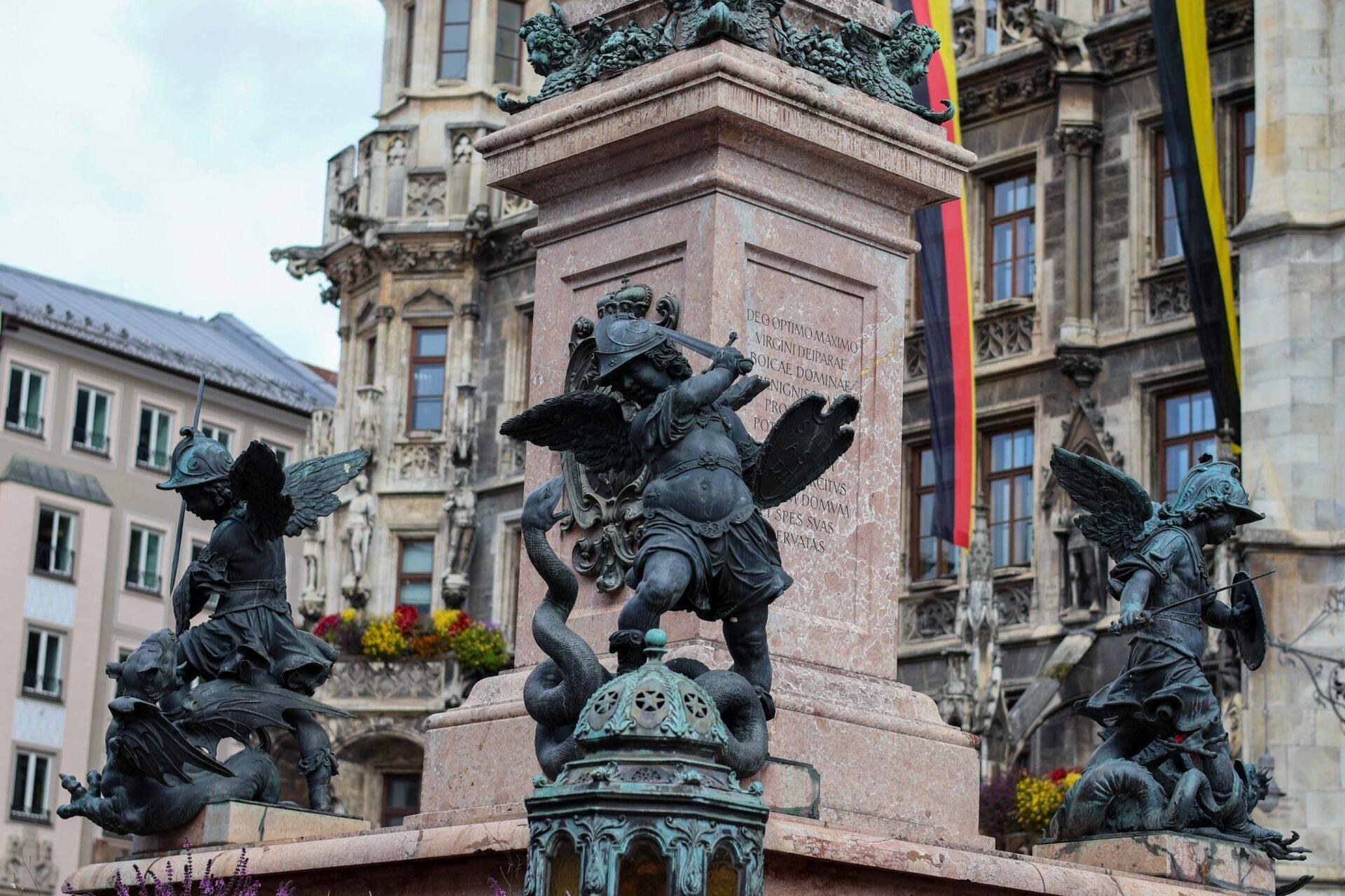 Monument avec statues d’anges ailés en bronze sur socle de pierre dans une place historique européenne entourée d’architecture classique
