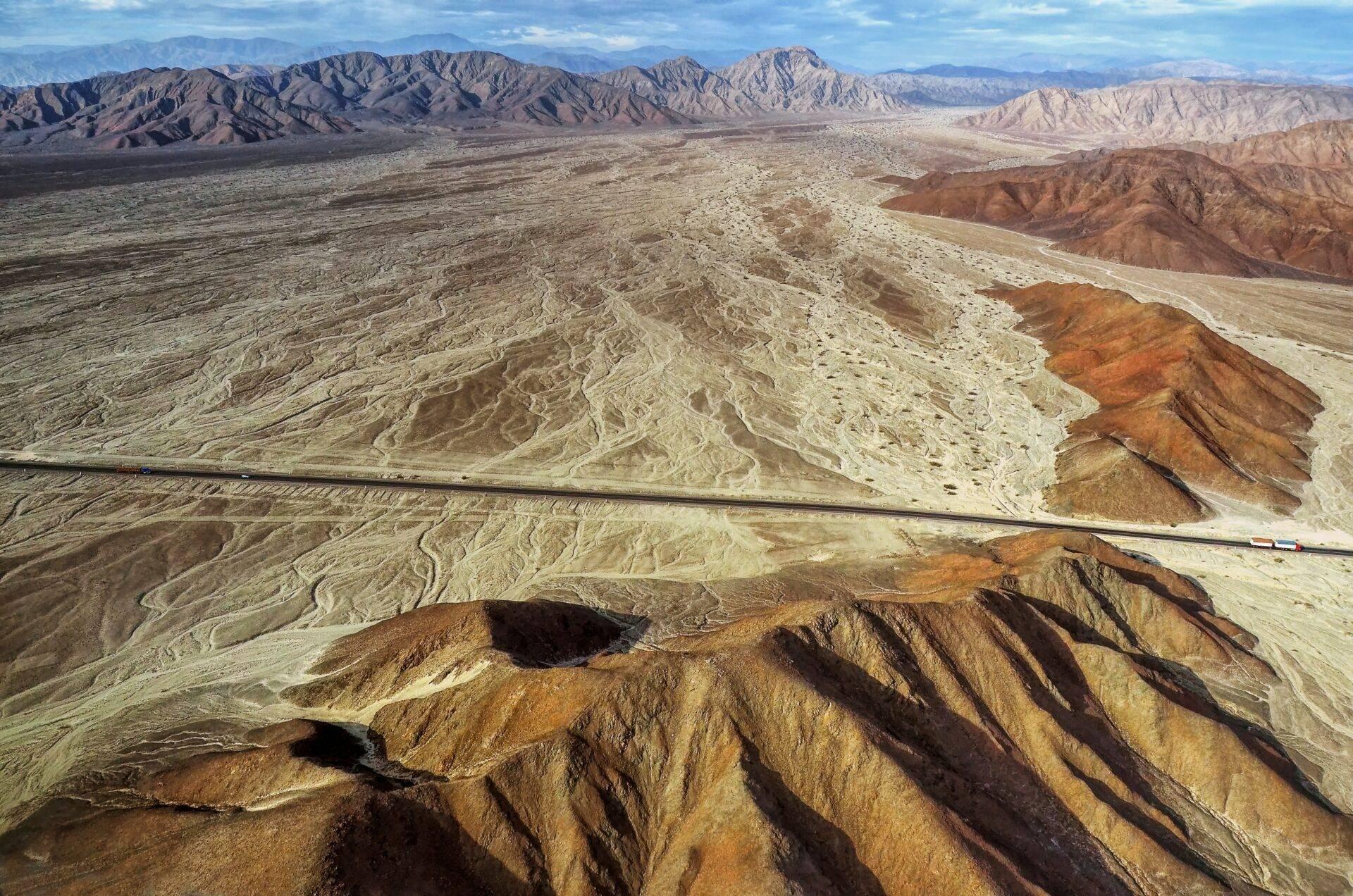 Vue aérienne d’une vallée désertique andine avec chaîne montagneuse, route rectiligne et formations géologiques colorées