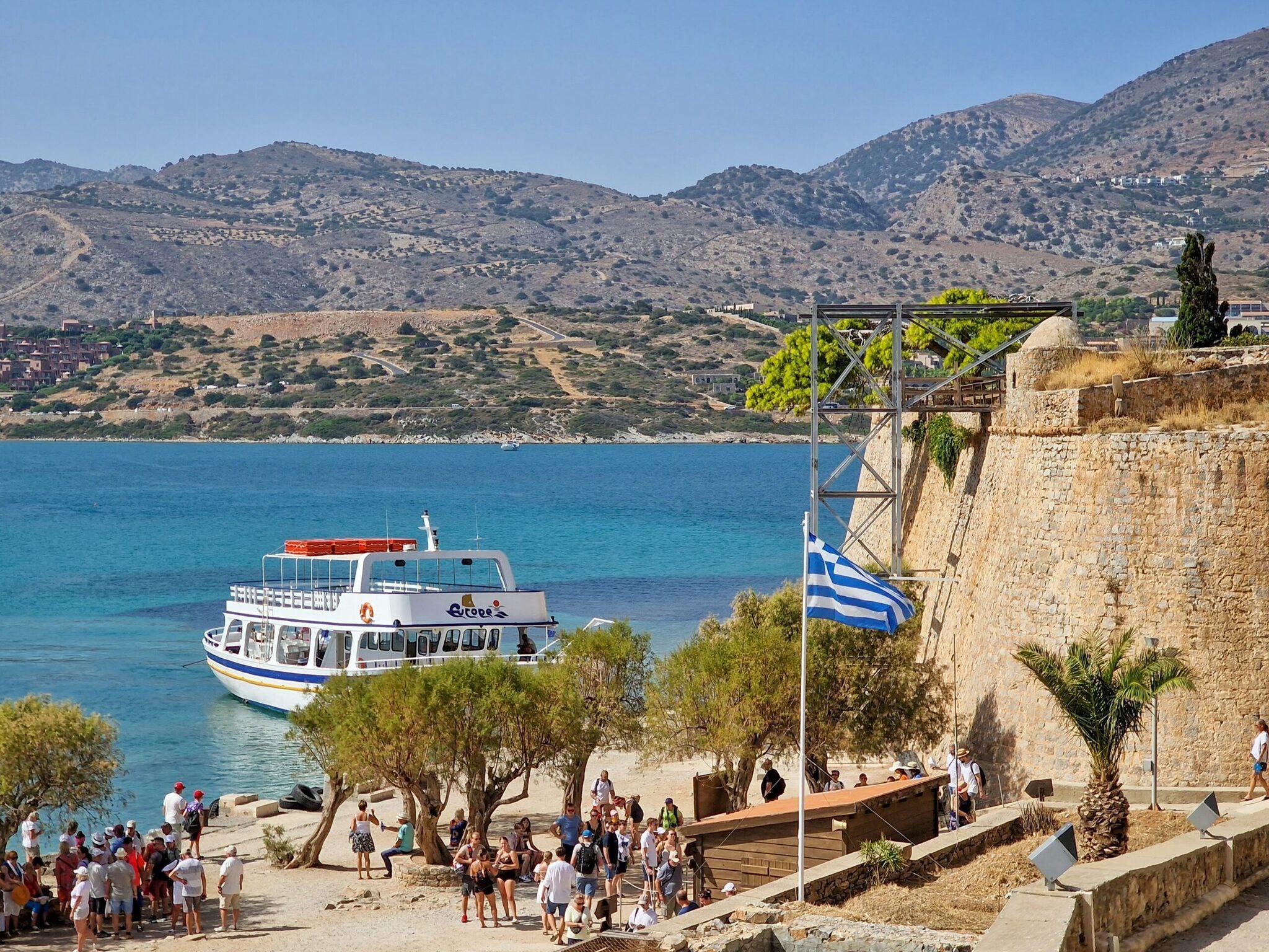 A white and blue tourist boat docked near a beach with a Greek flag and an arid mountain backdrop